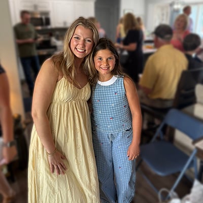Liv and Jenna stand together and pose for a photo at a graduation celebration. Other guests are gathered in the background inside a home.