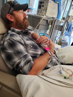  A seated adult holds a very small infant against their chest in a hospital room surrounded by medical equipment and monitoring lines.