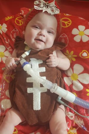 A young child with a tracheostomy sits on a red blanket wearing a brown football-themed outfit and a decorative headband.