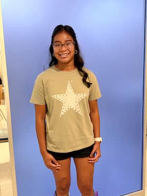 Karli standing against a blue backdrop wearing glasses, a light green T-shirt with a large star design, black shorts, and a wristwatch.