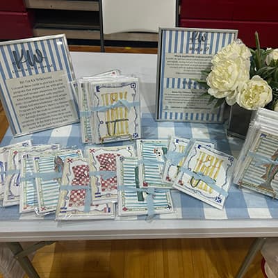 An assortment of greeting cards (sealed in plastic and tied with a light blue ribbon) are piled on a blue-striped cloth. The cards are watercolored with stripes and sailing flag motifs. Each features a different nautical subject: a life jacket, boat or fish. 