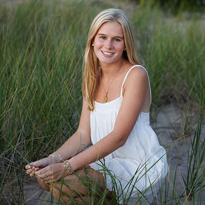 A young woman in a white eyelet dress and gold jewelry poses for a photo. She is kneeling in sand and seagrass and holding a blade of grass in her hand. 