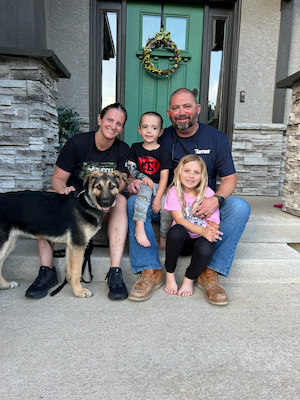 Koda and Family seated together on the front steps of a home with a dog, posing in front of a green front door with a decorative wreath.