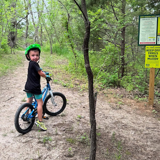 Koda wearing a bright green helmet standing with a blue bicycle on a wooded trail, looking back toward the camera near a posted trail information sign.