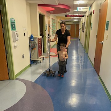 Caregiver assisting a child walking in a brightly lit hospital hallway, with medical equipment and patient room doors visible along the corridor.