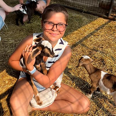 Matney sitting criss-cross in animal enclosure holding a brown and white baby goat.