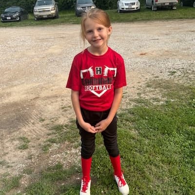 A young girl smiles wearing a red, black and white softball uniform in a gravel parking lot. Her shirt reads “H Cardinals Softball.”  
