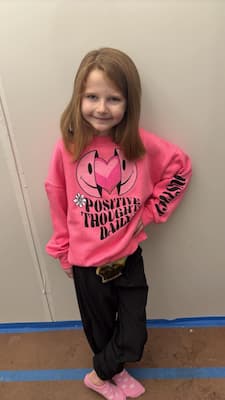 A young girl poses in a pink sweatshirt that reads “positive thoughts daily.” You can see a small medical pouch peeking out at her waistline.