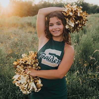 Teenager with wavy brown hair and braces poses in a green dance team uniform and gold metallic pom poms. Her top reads “Raiders” in front of a big letter “S.” She is standing in a field with the sun flaring behind her.