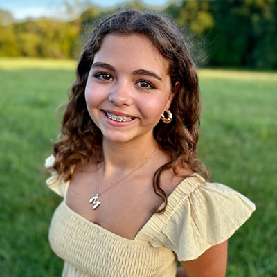 The photo is a portrait of a teenager wearing a yellow summer dress and smiling at the camera. She has brown, wavy hair and braces and is wearing a necklace with a gold letter “M” charm. A green field with grass and trees is out of focus behind her. 