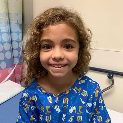 A young girl grins at the camera while sitting at the edge of a hospital bed. She has wavy light brown hair and is wearing a blue hospital gown with cartoon dogs and cats on it. 