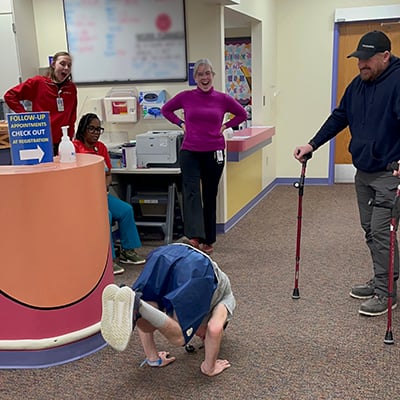 Several people look on with amazed expressions and smiles as a teen patient demonstrates his ability to do handstand pushups. The boy’s hands are on the floor and his legs are bent to hold his feet up off the ground as he lifts himself up and down.