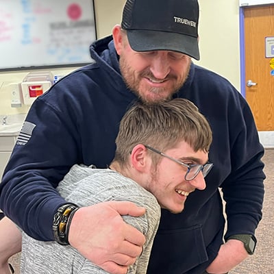 A dad in a navy blue sweatshirt and black ball cap wraps one arm around his teen son in a proud hug. Both dad and son have light brown hair and beards. The son is smiling and returning the hug.