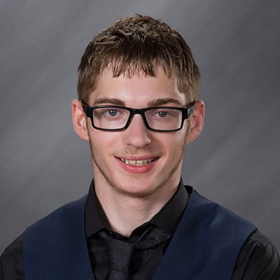 12th grade school photo of a young man with brown hair, black glasses and a slight beard. He wears a black dress shirt and black tie with a navy blue vest over it.