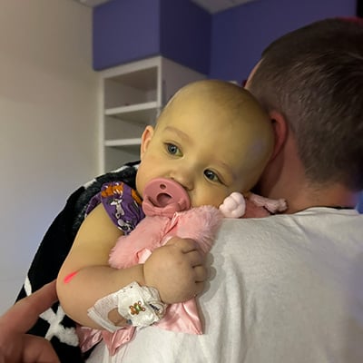 A man with his back to the camera holds a baby with an IV attached to her arm. Her skin is jaundiced, or appears distinctly yellow, and she’s sucking on a pink pacifier and holding a pink blanket.  