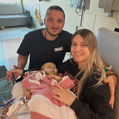 A couple in a hospital room, a man on the left and a woman on the right, smile at the camera while holding a small baby girl. The baby has tubes coming from her face and body, which is mostly covered by a pink and white striped blanket. The tape holding tubes to the baby’s nose has “I am so brave!” written on it.  