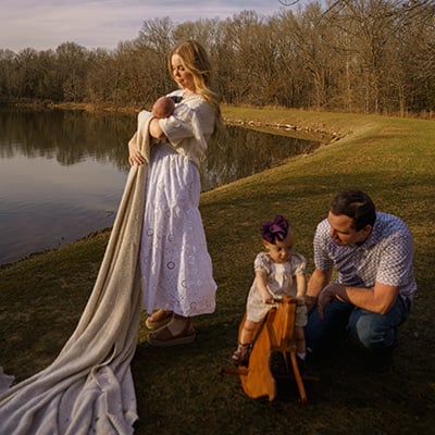 A family poses on a lakeshore. The mom is standing, wearing a white dress and holding a baby wrapped in a long blanket that is trailing in the foreground. A toddler is on a wooden rocking horse to the mom’s right, and the dad is crouched down beside her. 