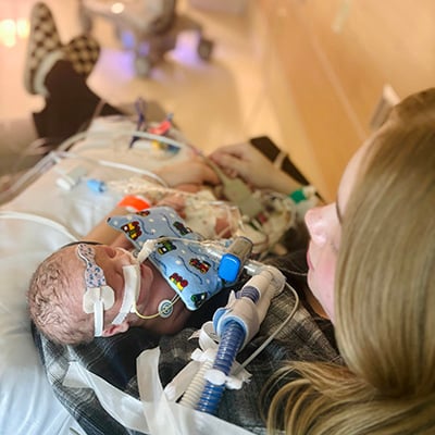 A mom holds a baby in a hospital room. The baby has many wires and tubes attached to his small body, and there is more hospital equipment blurred in the background. 