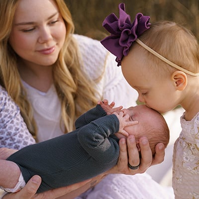 A toddler kisses her baby brother on the forehead. He’s being held up by his dad, who is mostly out of frame, while mom looks on behind them. The toddler is wearing a headband with a big purple bow.