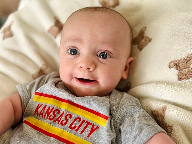 A baby with big blue eyes smiles at the camera. He is wearing a onesie that reads “Kansas City” and is laying on a cream blanket with teddy bear illustrations. 