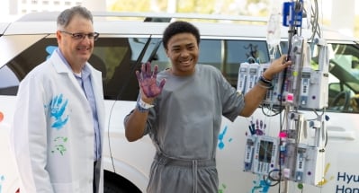 Patient standing beside a medical infusion pole outdoors, raising a hand covered in purple paint next to a clinician in a white coat with a blue handprint. A white vehicle is visible in the background.