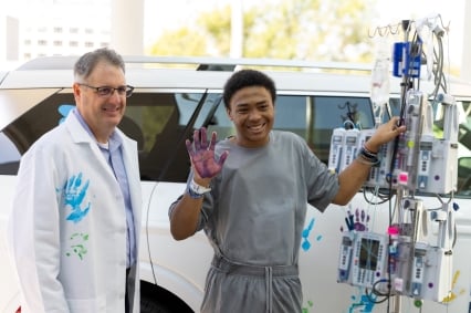 Patient standing beside a medical infusion pole outdoors, raising a hand covered in purple paint next to a clinician in a white coat with a blue handprint. A white vehicle is visible in the background.