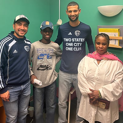 A patient stands in a clinic exam room with several visitors wearing soccer apparel, positioned together for a photo during a hospital visit.