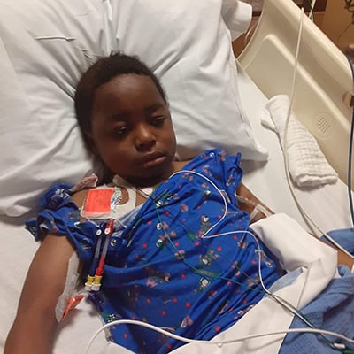 A patient lies in a hospital bed wearing a patterned hospital gown, with medical tubing and monitoring leads visible and connected to equipment beside the bed.