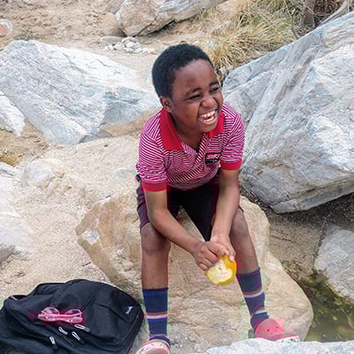 A young person sits on a large rock in an outdoor, rocky area, holding an orange and wearing a red striped shirt, with a backpack placed on the ground nearby.