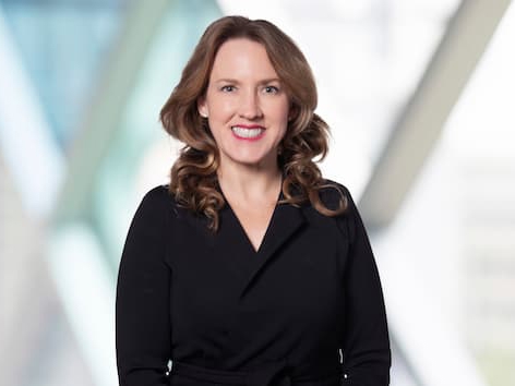Professional headshot of a person with shoulder-length wavy hair wearing a dark blazer, seated in front of a softly blurred office background with geometric glass and metal architectural elements.