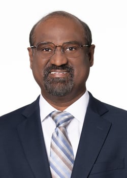 A man with a goatee and glasses is wearing a white, collared shirt and tie with a dark blue blazer.