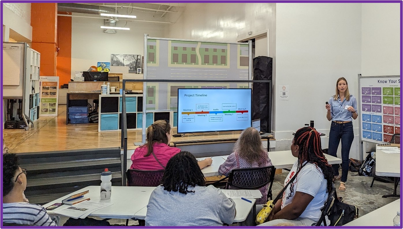 Community members discussing environmental conditions around bus stops with Drs. Grimes and Carlson. They sit in a room at rows of tables. There is a screen behind the presenters.