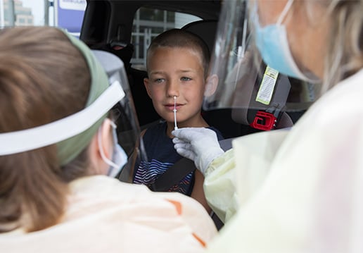 Two nurses in PPE performing a nasal swab test on boy in a car