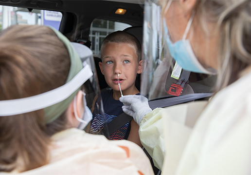 Two nurses in PPE performing a nasal swab test on boy in a car