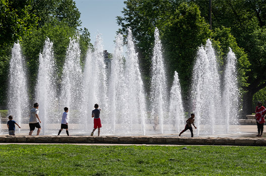Photo of five children playing near fountains with an adult standing nearby.