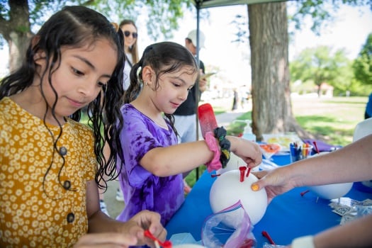 Two girls work on a craft project at a table in a park. One girl is holding a red popsicle.