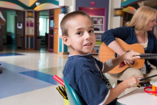 A young boy tips back in his chair as a woman plays guitar in a school environment.