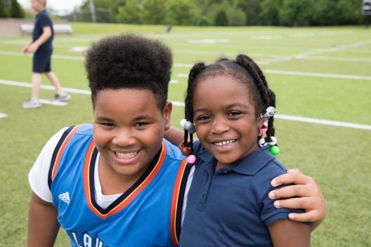A young Black boy and Black girl with braids stand on a football field with arms around each other.
