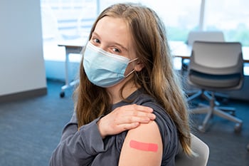 Girl wearing a face mask and showing off the pink adhesive bandage on her arm after receiving a vaccination at Children's Mercy.